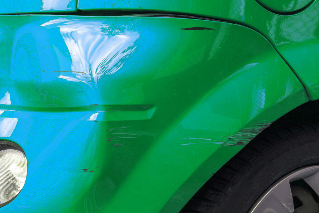 Nahaufnahme einer grünen Autoecke mit Kratzern, Delle und sichtbarem Vorderrad