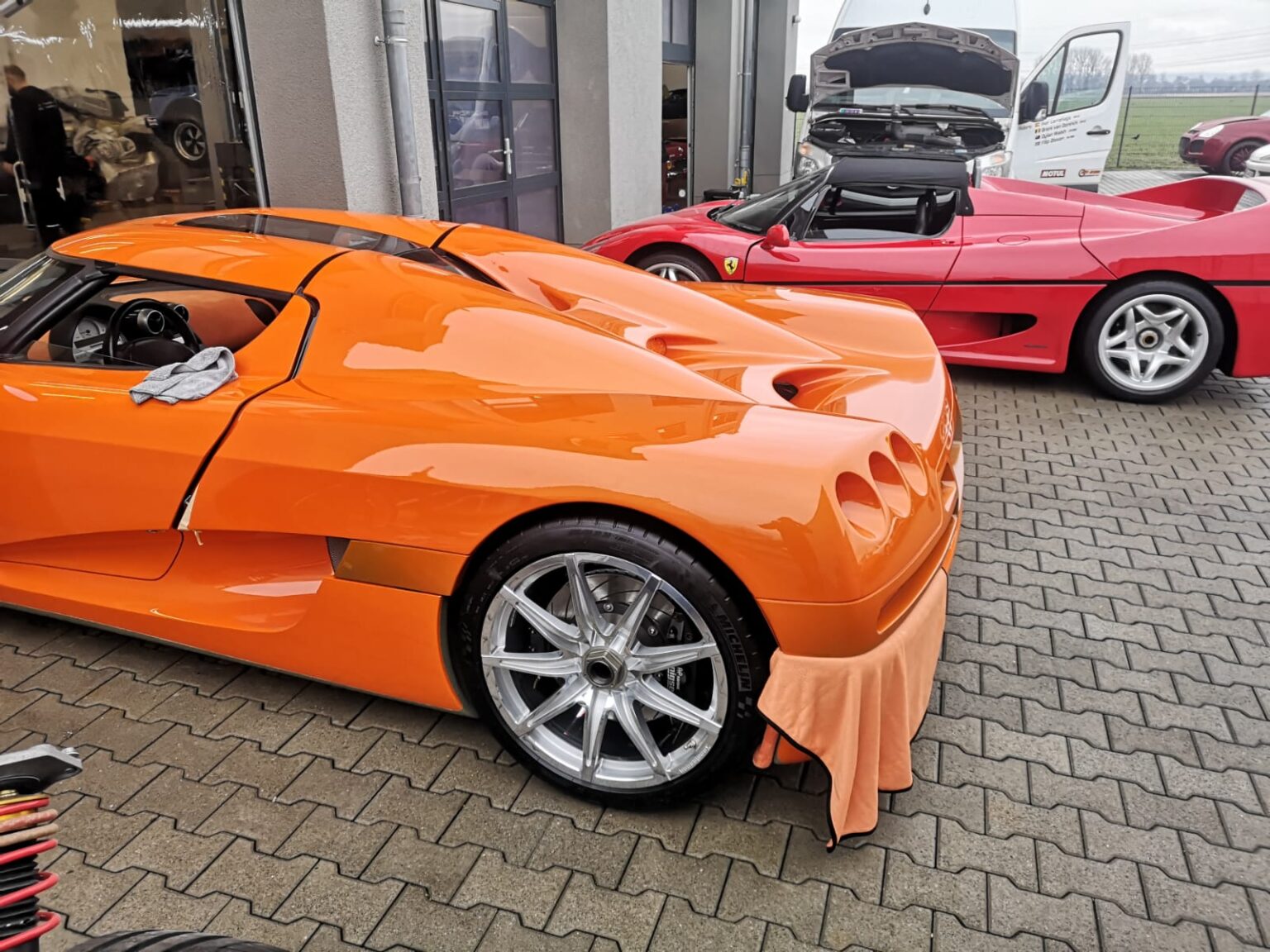 Orange Sportwagen und roter Ferrari stehen vor einer Werkstatt; beim orangen Auto hängt ein Tuch am Kotflügel
