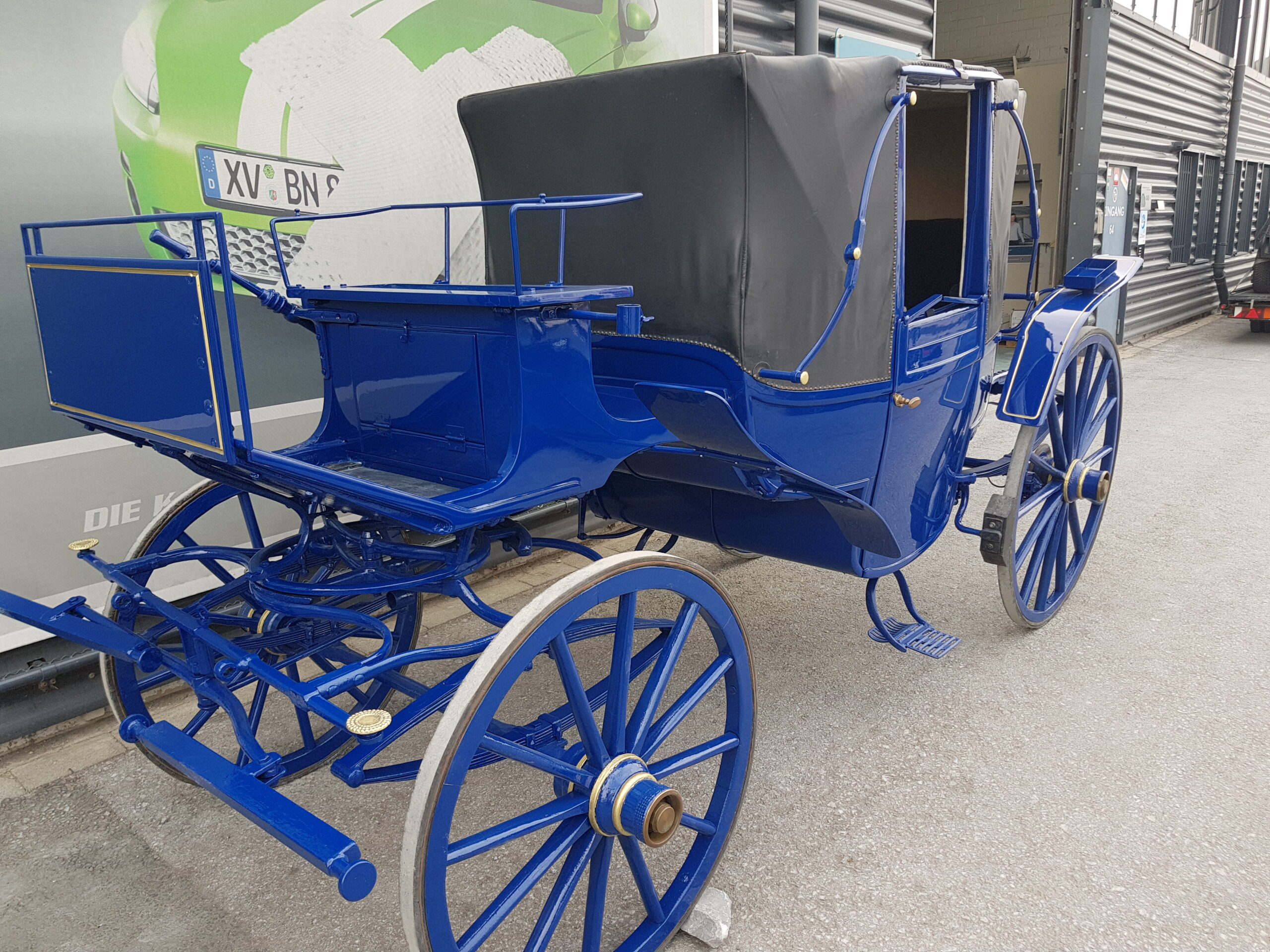 Blauer historischer Pferdewagen mit schwarzer Abdeckung und großen Speichenrädern vor einer Wandgrafik