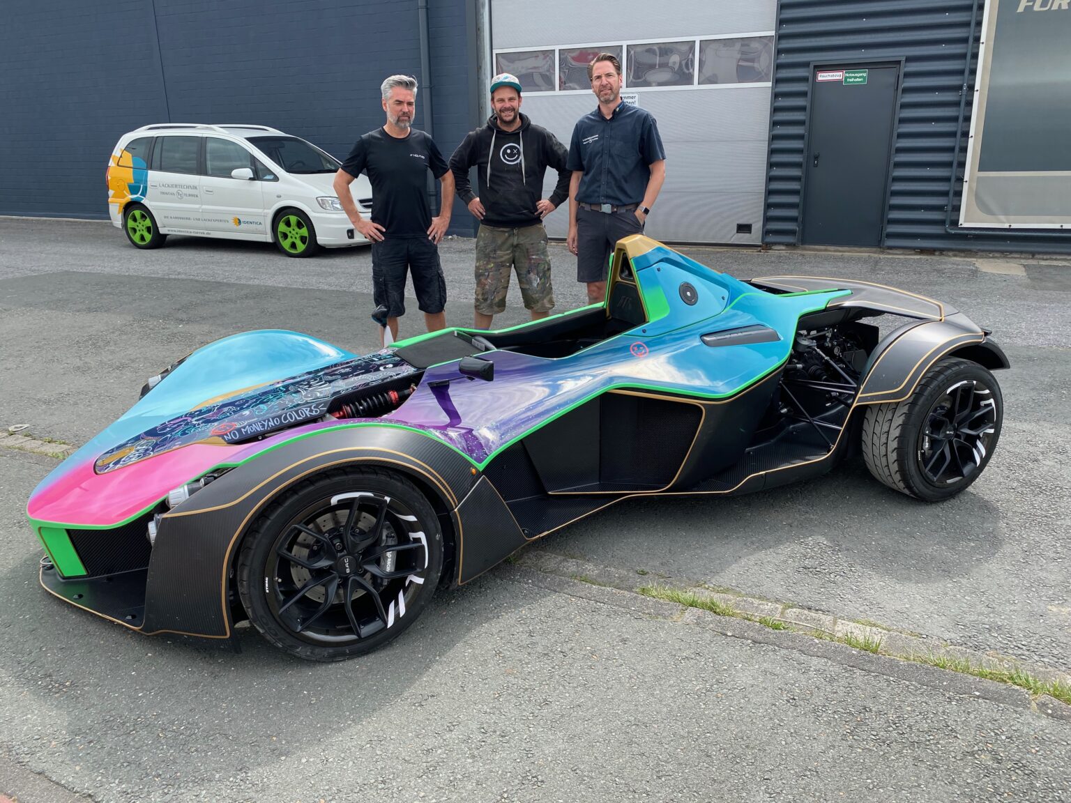 Drei Männer stehen vor einem bunt lackierten, offenen Sportwagen mit futuristischem Design