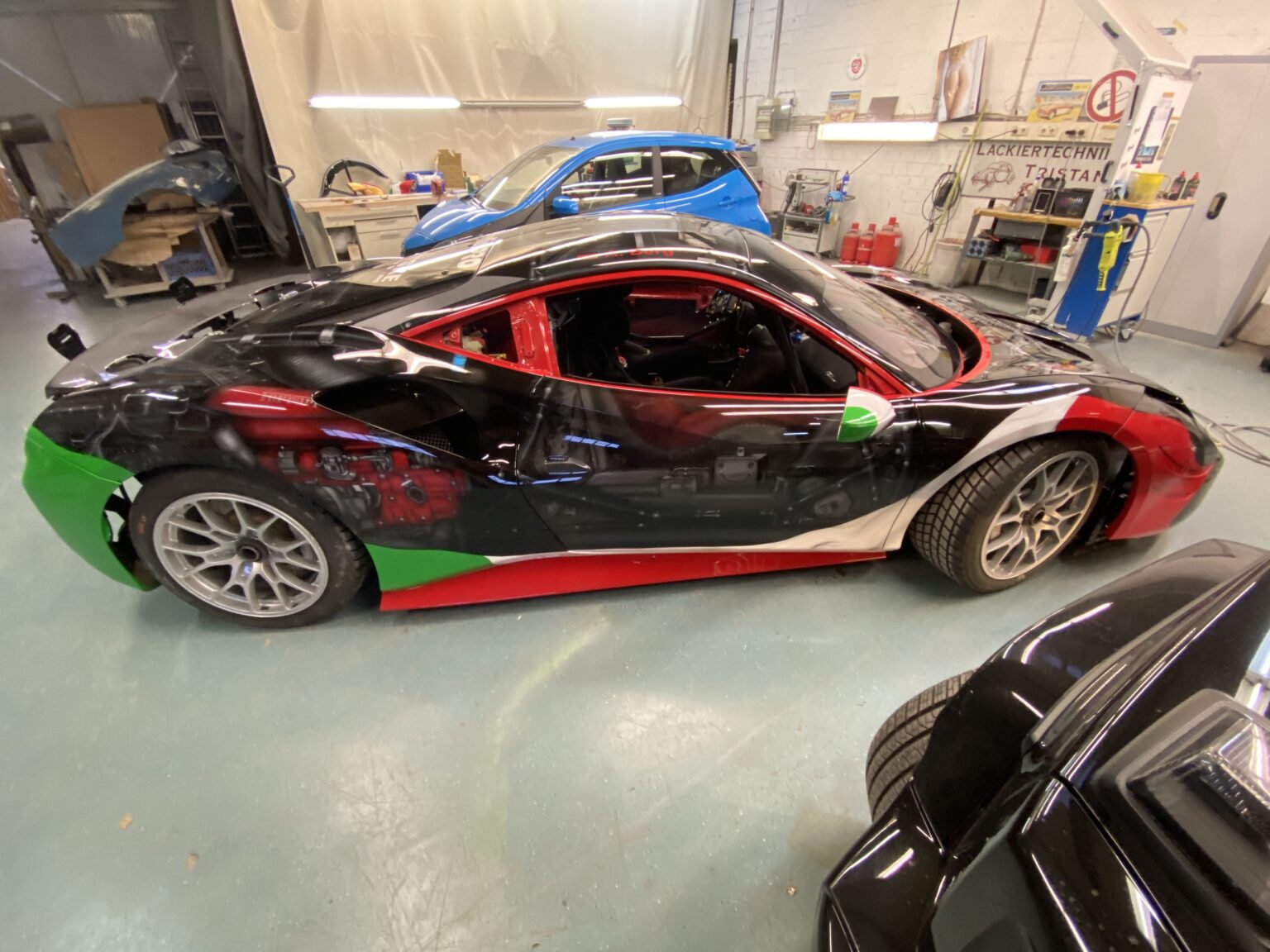 Sportwagen mit schwarz-glänzender Lackierung und rot-grün-weißen Details in einer Werkstatt, daneben ein blauer Kleinwagen