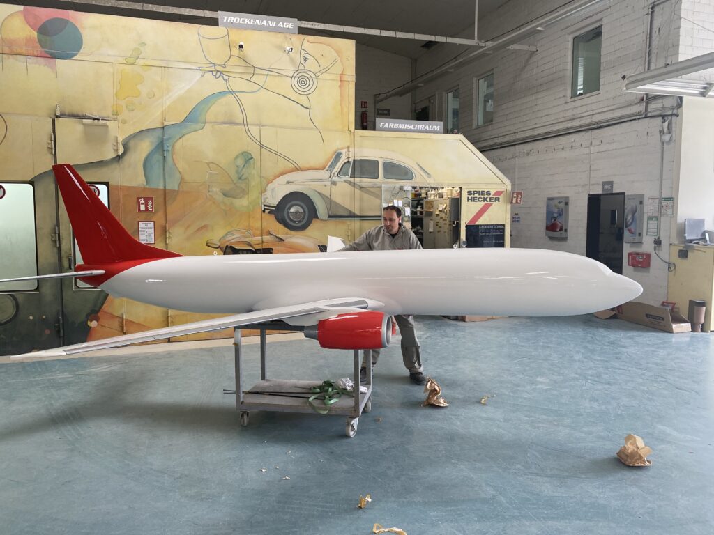 Ein kleines Flugzeugrumpfmodell in einer Lackierwerkstatt, daneben ein Mann bei der Arbeit
