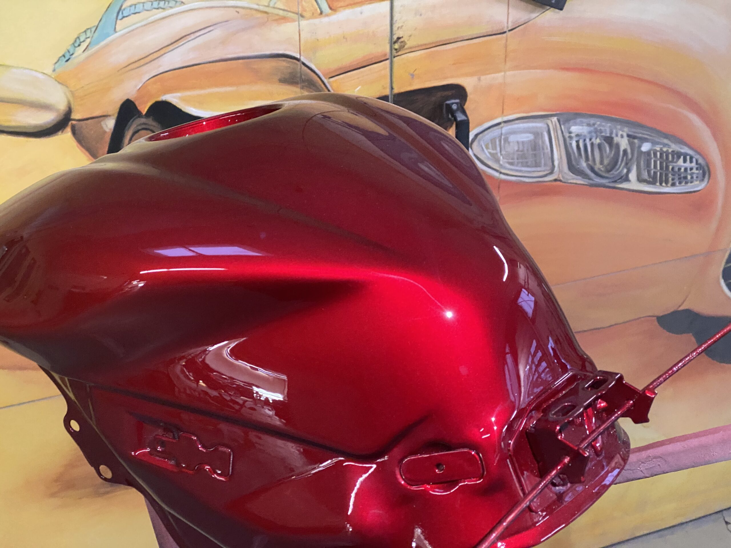 Glänzend roter Motorrad-Tank vor einer gelb-orange bemalten Wand mit großem Fahrzeuggesicht im Hintergrund