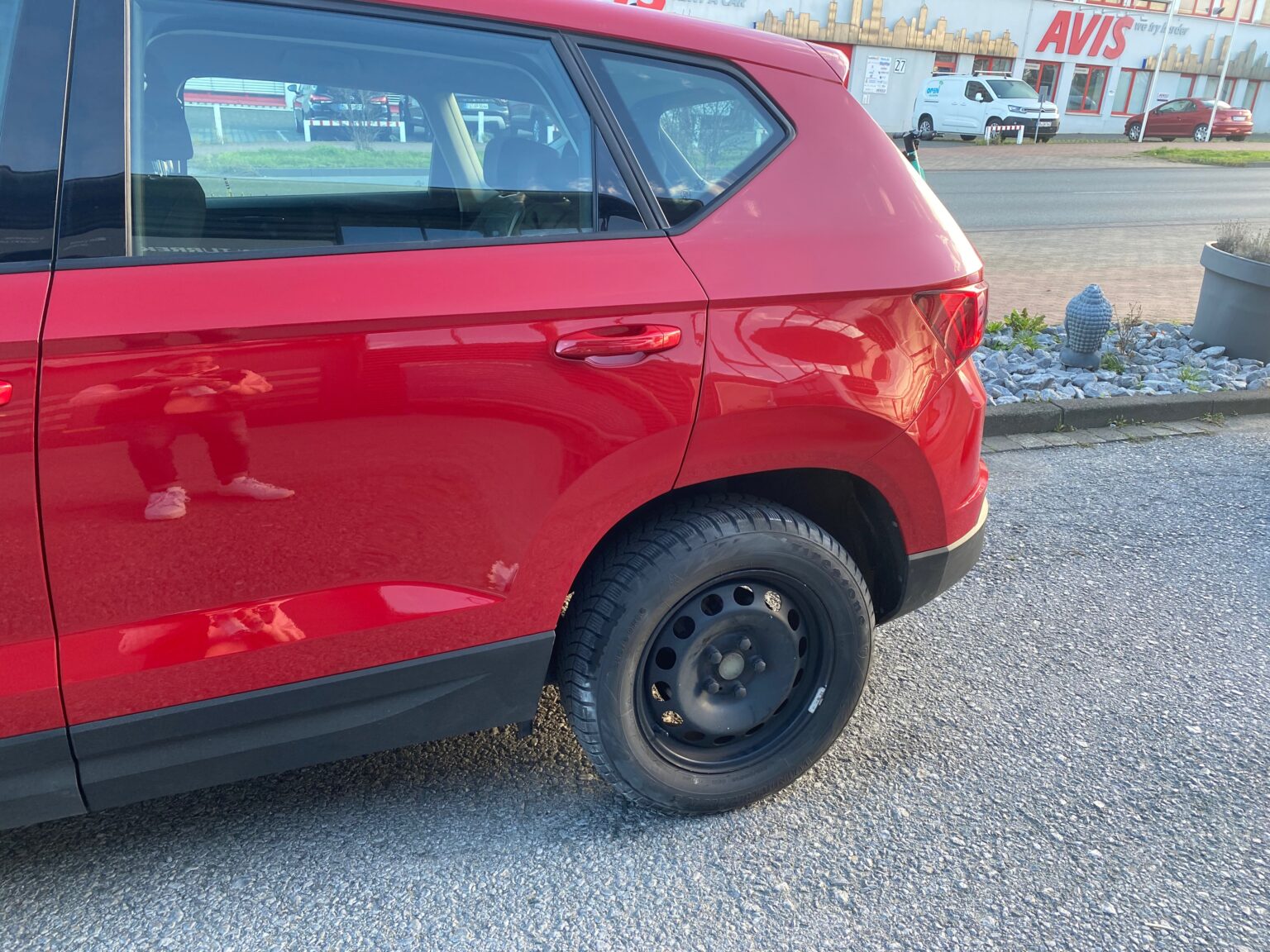 Rote Autoseite mit schwarzem Stahlrad auf einem Parkplatz vor einer AVIS-Filiale