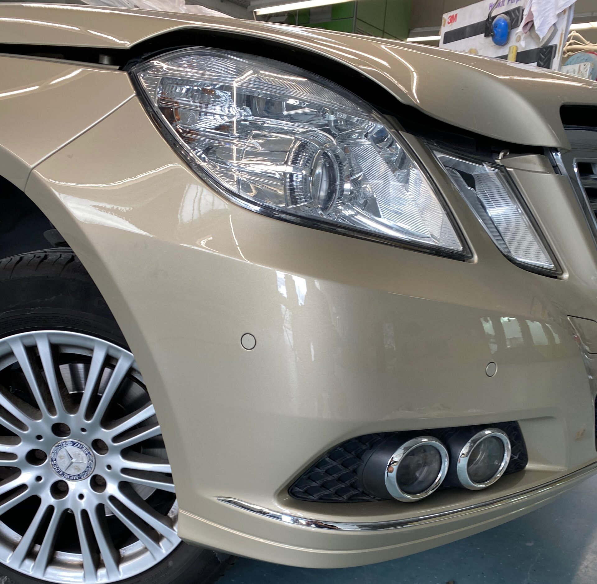 Nahaufnahme der Front eines beige-goldenen Mercedes mit Scheinwerfer, Felge und zwei runden Auspuffblenden
