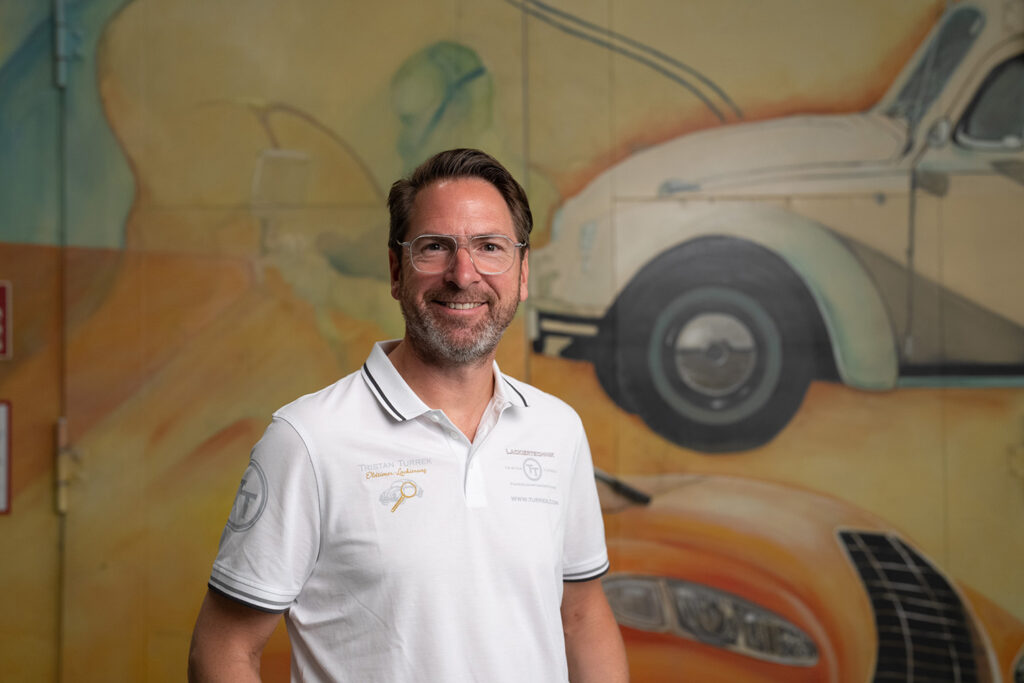 Lächelnder Mann in weißem Poloshirt vor einer Wandmalerei mit Auto