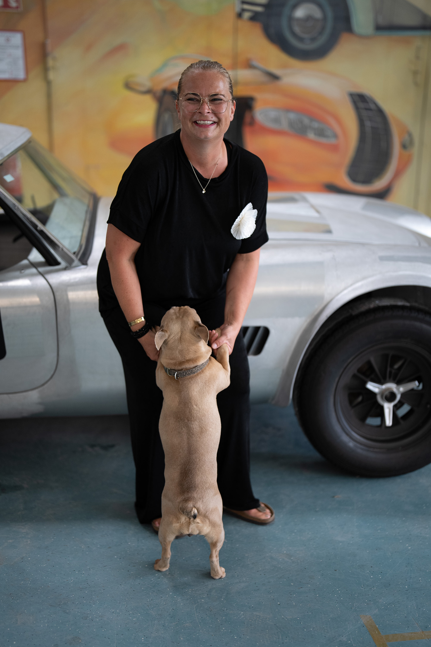 Lächelnde Frau im schwarzen Outfit mit Mops vor einem silbernen Auto und einer Wandmalerei im Hintergrund